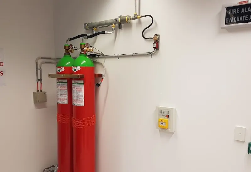 Two red and green fire suppression cylinders mounted on a wall with connected piping and a manual release switch in a clean equipment room.