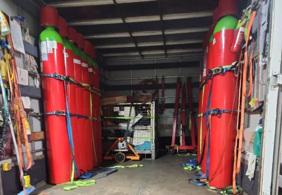 Several large red and green gas cylinders securely strapped upright inside the back of an Isuzu truck, with tie-down straps and equipment organized along the walls and floor, ready for transport or delivery.