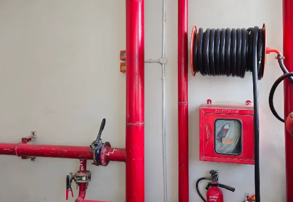 Fire safety system setup featuring red water pipes, a mounted fire hose reel, a fire extinguisher, and a wall cabinet containing a coiled hose, designed for emergency firefighting and suppression.