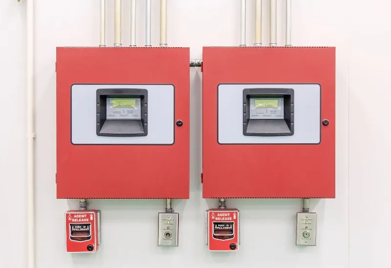 Two red fire alarm control panels mounted on a wall, each with a display screen and connected manual pull stations below, used for fire detection and suppression system monitoring.