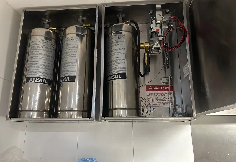 Close-up of a stainless steel Ansul fire suppression system with three pressurised cylinders, hoses, and activation controls, mounted on a commercial kitchen wall for automatic fire protection.