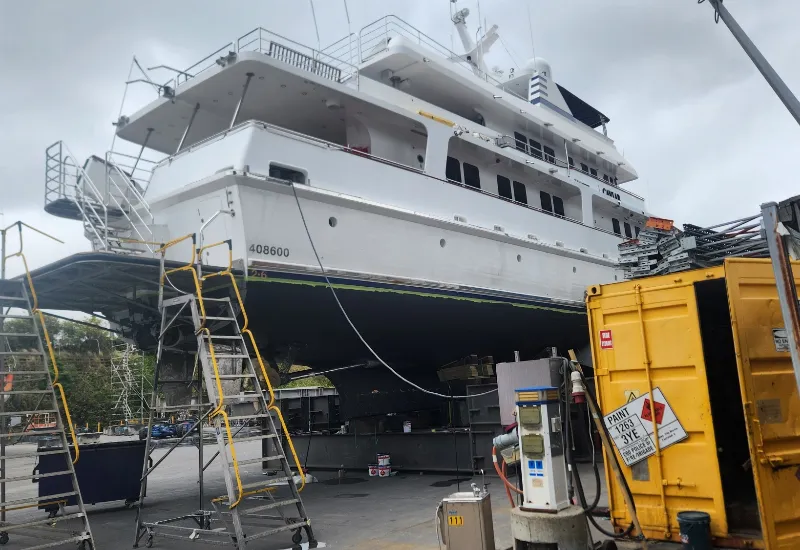 A large white yacht elevated on supports in a shipyard, surrounded by scaffolding and maintenance equipment, with a yellow container and fuel pump in the foreground under a cloudy sky