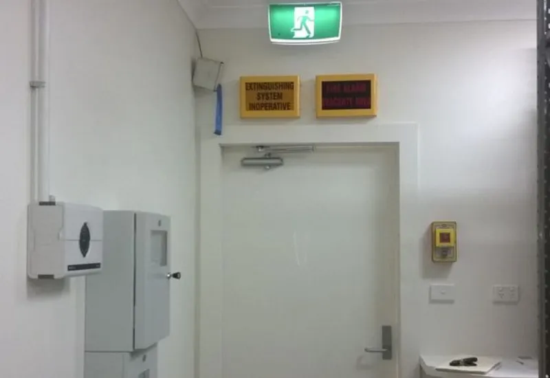 White hallway with an emergency exit door, green exit sign, and fire safety warning signs above the door