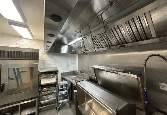 Commercial kitchen setup with stainless steel appliances, overhead ventilation hoods, industrial oven, and deep sink, designed for high-efficiency cooking and equipped for professional fire safety and hygiene standards.