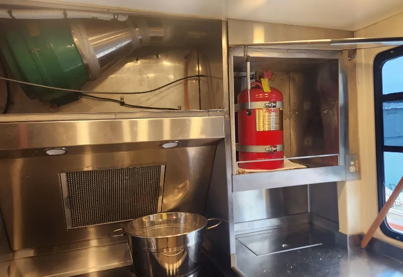 Commercial kitchen setup showing a stainless steel cooking area with ventilation ducting, a large cooking pot on the stove, and a mounted red Fireboy fire suppression system for safety.