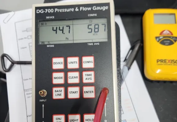 Close-up of a DG-700 Pressure and Flow Gauge device used for fan integrity testing, displaying readings of 44.7 Pa and 587 m³/h. The instrument, labeled “The Energy Conservatory” and connected with red and clear tubing, rests on top of a Minneapolis Blower Door unit beside papers and a yellow measuring device.