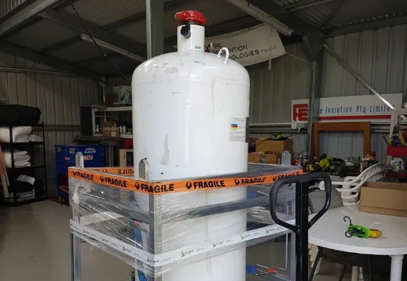 A large white industrial gas cylinder secured on a metal frame with orange “Fragile” tape inside a workshop or storage shed, surrounded by tools, boxes, and equipment.