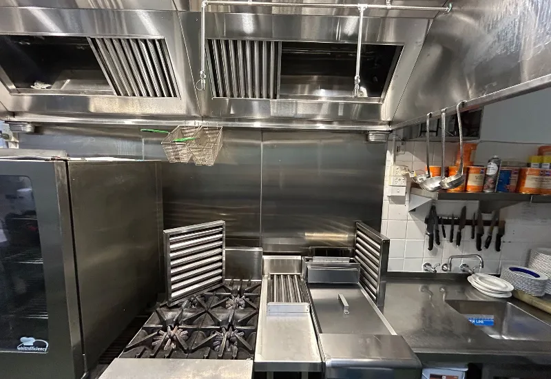 Professional commercial kitchen with stainless steel appliances, gas burners, deep fryer, grill, and overhead exhaust hood, designed for efficient cooking and equipped with ventilation and utensil storage.