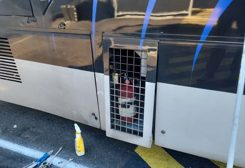 A fire extinguisher secured behind a metal grate on the side panel of a vehicle, with tools and a spray bottle on the ground nearby.