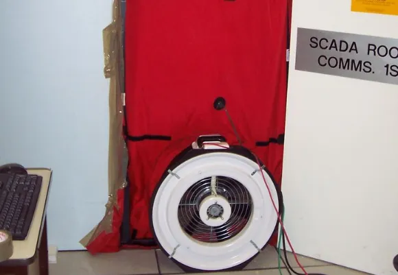 A blower door fan setup installed in a doorway with a red air-sealing panel, used for air pressure and integrity testing in a building. The door sign reads “SCADA Room COMMS. 1S,” and computer equipment is visible nearby.